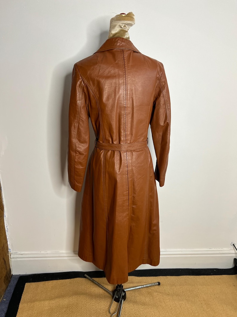 May include: A vintage brown leather trench coat with a belted waist and long sleeves. The coat is displayed on a mannequin, showcasing its tailored fit and classic design. The coat's color is a warm, rich brown.