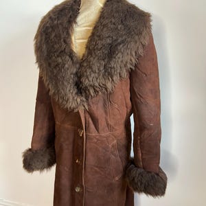 May include: A vintage brown suede coat with a faux fur collar and cuffs. The long coat has a button-down front and is displayed on a mannequin. The coat is a medium brown color.