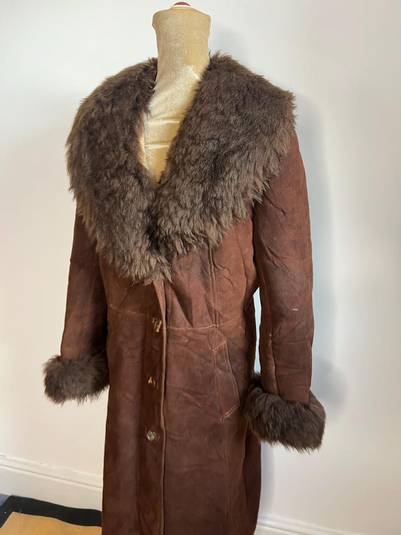 May include: A vintage brown suede coat with a faux fur collar and cuffs. The long coat has a button-down front and is displayed on a mannequin. The coat is a medium brown color.