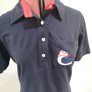 May include: A navy blue polo shirt with a red collar and a white button placket. The shirt has a pocket on the left side with a sequined penguin design.