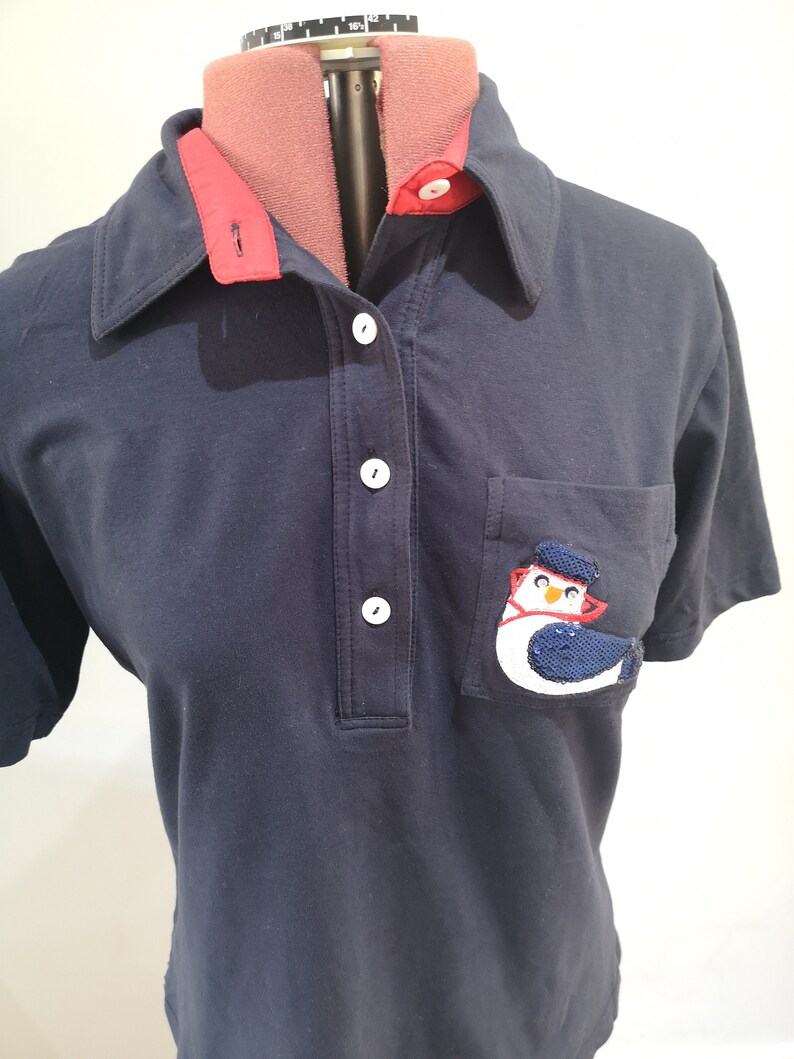May include: A navy blue polo shirt with a red collar and a white button placket. The shirt has a pocket on the left side with a sequined penguin design.