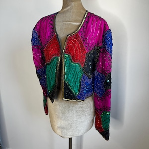 May include: A colorful, vintage jacket with a patchwork design. The jacket features a variety of colors, including pink, red, green, blue, and black, and is embellished with sequins and beads. The jacket is displayed on a mannequin.