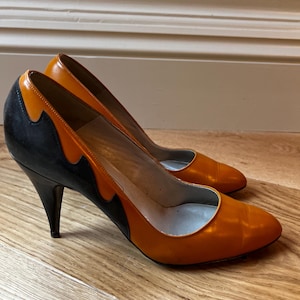Könnte beinhalten: Ein Paar orangefarbene und schwarze High Heels. Die Schuhe haben eine spitze Zehenpartie und einen schwarzen Absatz. Der obere Teil des Schuhs ist orange, mit einem schwarzen Akzent an der Seite. Die Schuhe sind aus Leder.