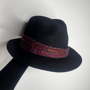 Pode incluir: Um chapéu fedora preto com uma faixa estampada em vermelho, azul e dourado. O chapéu tem uma aba larga e um design clássico. A faixa apresenta um intrincado padrão paisley.