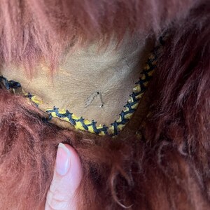 May include: Close-up of a section of a brown, shaggy fur item. The fur is a deep reddish-brown color. The item has a tan leather interior with yellow and black stitching along the edge. A finger is visible in the image.