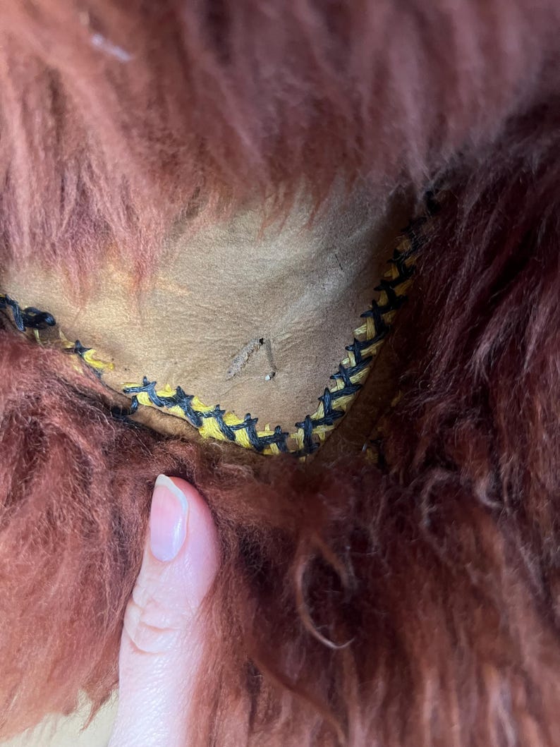 May include: Close-up of a section of a brown, shaggy fur item. The fur is a deep reddish-brown color. The item has a tan leather interior with yellow and black stitching along the edge. A finger is visible in the image.