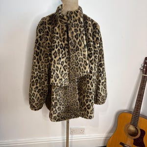 Op de afbeelding: Een jas van imitatiebont met lange mouwen en een luipaardprint in bruin, zwart en beige. De jas heeft een hoge kraag en een bijpassend vastzittend sjaaldetail dat aan de voorzijde naar beneden hangt. Het kledingstuk wordt getoond op een paspop.