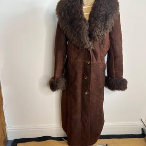 May include: A long, brown suede coat with a large, dark brown fur collar and cuffs. The coat has three buttons down the front and is displayed on a mannequin. The coat is a vintage style.