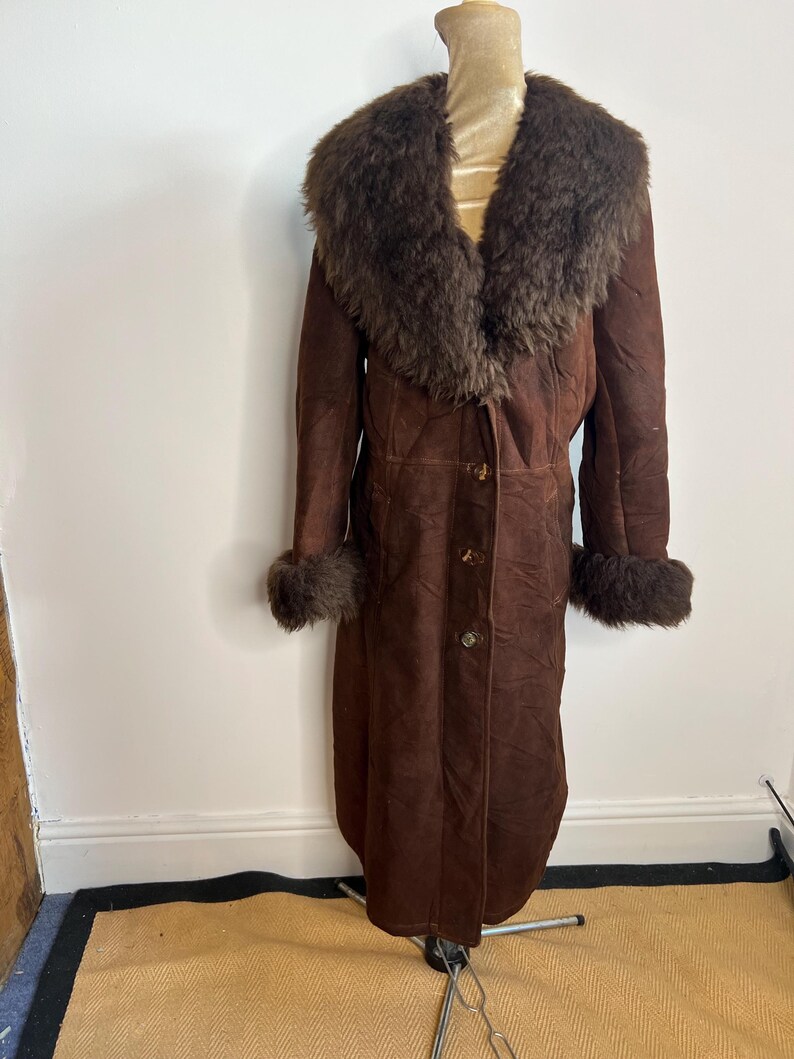 May include: A long, brown suede coat with a large, dark brown fur collar and cuffs. The coat has three buttons down the front and is displayed on a mannequin. The coat is a vintage style.