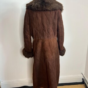 May include: A long, brown shearling coat with a dark brown fur collar and cuffs. The coat is displayed on a mannequin and has a classic, vintage style. The coat is made of a soft, suede-like material.
