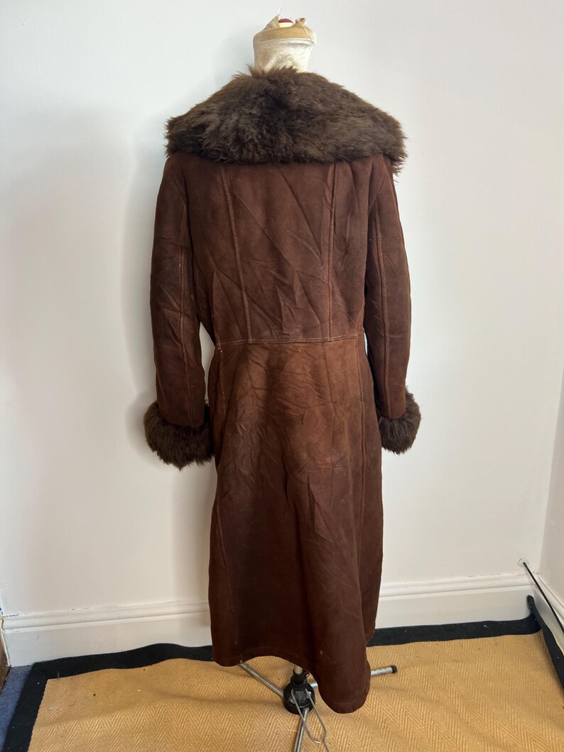 May include: A long, brown shearling coat with a dark brown fur collar and cuffs. The coat is displayed on a mannequin and has a classic, vintage style. The coat is made of a soft, suede-like material.