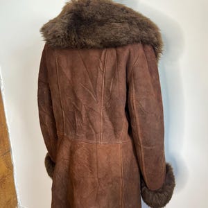 May include: A vintage brown suede coat with a fur collar and cuffs. The long coat is displayed on a mannequin, showcasing its full length and design. The coat features a classic silhouette with a tailored fit.