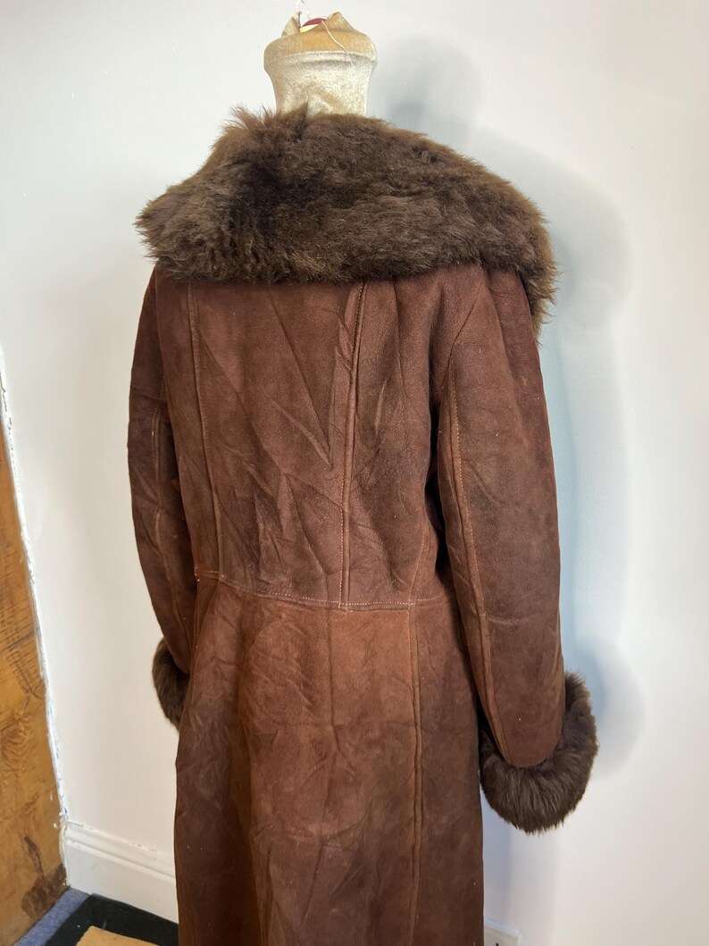 May include: A vintage brown suede coat with a fur collar and cuffs. The long coat is displayed on a mannequin, showcasing its full length and design. The coat features a classic silhouette with a tailored fit.