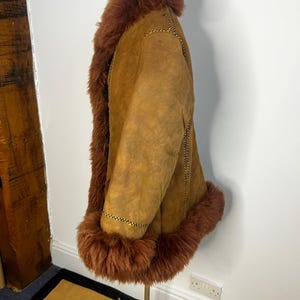 May include: A tan suede coat with brown faux fur trim around the collar, cuffs, and hem. The coat has a relaxed fit and features decorative stitching along the seams. The coat is displayed on a mannequin.