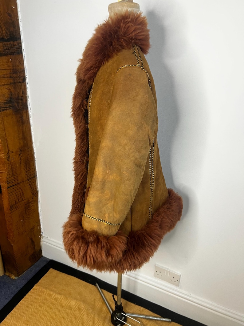 May include: A tan suede coat with brown faux fur trim around the collar, cuffs, and hem. The coat has a relaxed fit and features decorative stitching along the seams. The coat is displayed on a mannequin.
