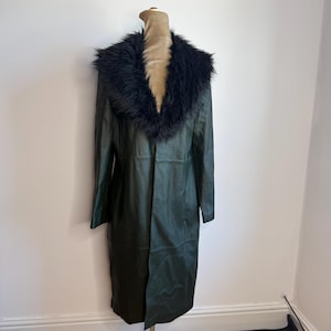 May include: A dark green leather coat with a black faux fur collar is displayed on a mannequin. The coat is long, reaching to the mid-calf, and has a classic, tailored design. The mannequin is on a metal stand.
