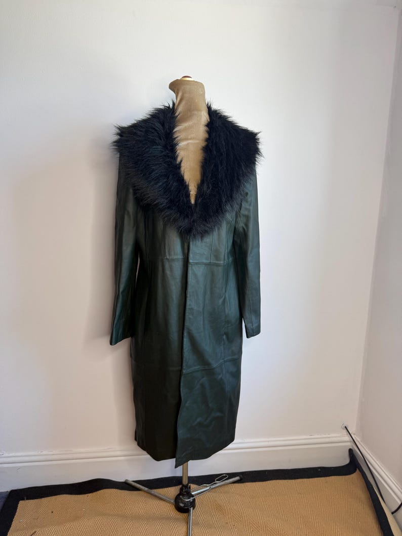 May include: A dark green leather coat with a black faux fur collar is displayed on a mannequin. The coat is long, reaching to the mid-calf, and has a classic, tailored design. The mannequin is on a metal stand.