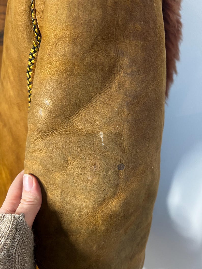 May include: Close-up of a brown leather jacket sleeve. The jacket has a distressed appearance with visible wear and tear. A black and yellow braided cord detail runs along the edge. The interior lining is a reddish-brown faux fur.