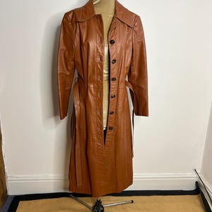 May include: A vintage, long, brown leather coat with a button-up front and a matching belt. The coat is displayed on a mannequin and has a classic, tailored design. The coat's color is a warm, medium brown, and it appears to be in good condition.
