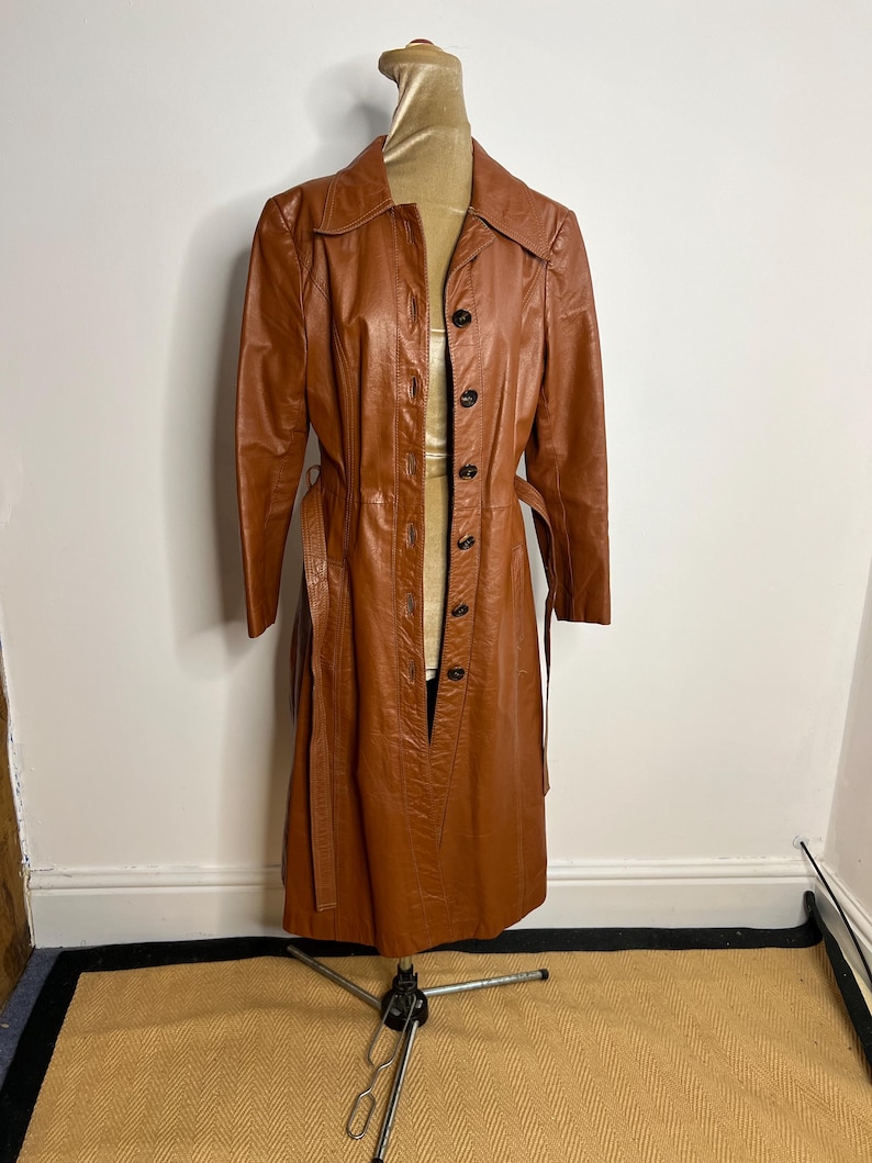 May include: A vintage, long, brown leather coat with a button-up front and a matching belt. The coat is displayed on a mannequin and has a classic, tailored design. The coat's color is a warm, medium brown, and it appears to be in good condition.