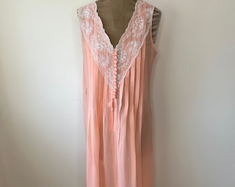May include: A peach-colored vintage nightgown with a V-neckline and delicate white lace trim. The sleeveless gown features vertical pleats and small peach-colored buttons down the front. The hemline is also trimmed with white lace.