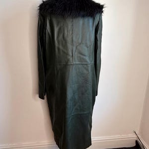 May include: A long, dark green leather coat with a black faux fur collar. The coat is displayed on a mannequin, showcasing its full length and tailored fit. The coat has a classic design, suitable for various occasions.
