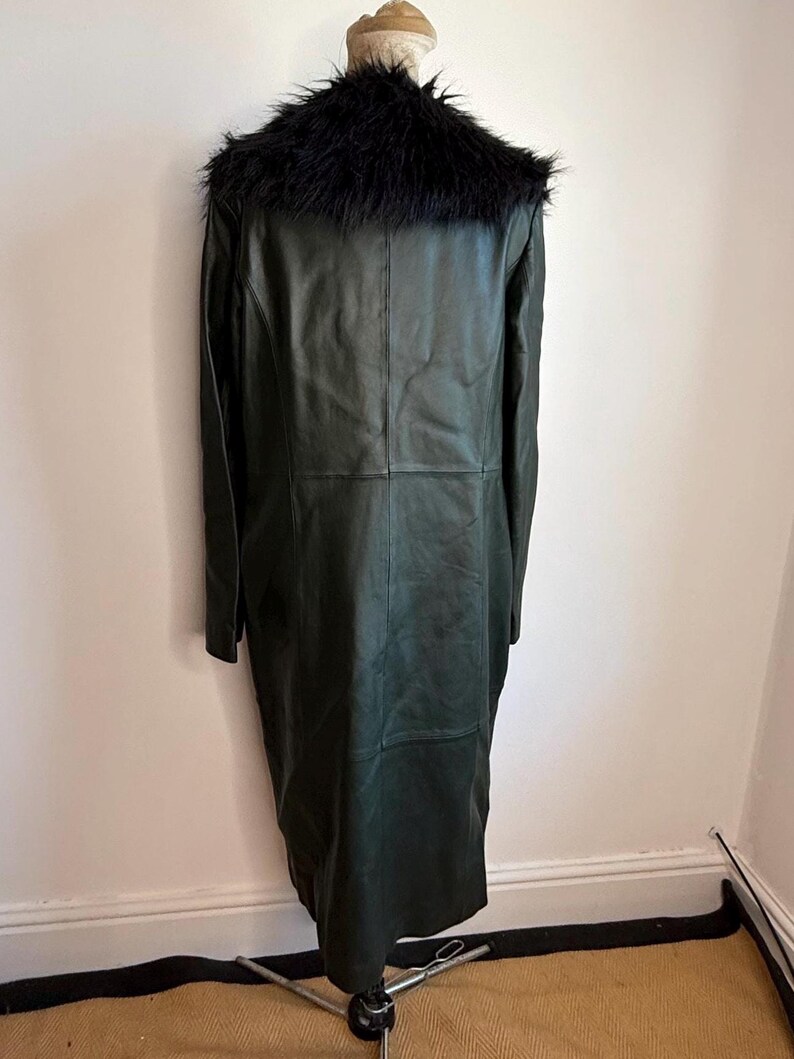 May include: A long, dark green leather coat with a black faux fur collar. The coat is displayed on a mannequin, showcasing its full length and tailored fit. The coat has a classic design, suitable for various occasions.