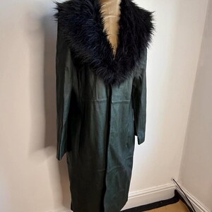 May include: A dark green, full-length coat with a black faux fur collar. The coat is displayed on a mannequin. The coat appears to be made of leather or a leather-like material. The coat has a classic, timeless design.
