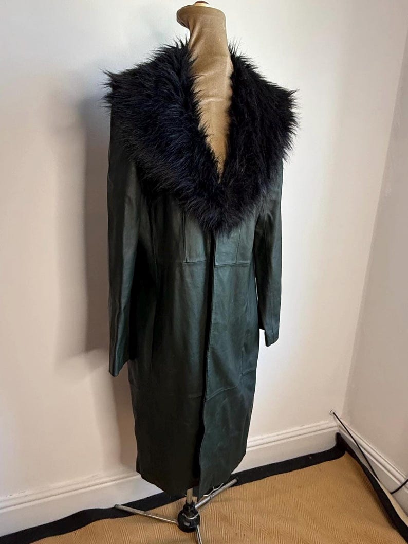 May include: A dark green, full-length coat with a black faux fur collar. The coat is displayed on a mannequin. The coat appears to be made of leather or a leather-like material. The coat has a classic, timeless design.