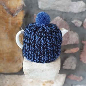 Könnte beinhalten: Blaue und schwarze gestrickte Teekannen-Cosy mit einem blauen Bommel oben. Die Cosy liegt auf einer weißen Steinoberfläche.