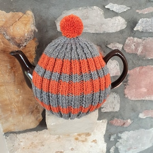 May include: A grey and orange striped knitted teapot cosy with a pom pom on top. The cosy is on a brown teapot.