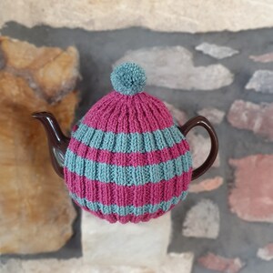 May include: A brown teapot is covered by a knitted tea cosy. The cosy has magenta and teal horizontal stripes, and a teal pom-pom. The cosy is designed to keep the tea warm, and measures approximately 20cm in diameter.