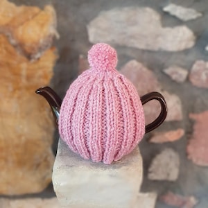 May include: A pink knitted tea cosy with a pom-pom on top, covering a brown teapot. The cosy has a ribbed design and is sitting on a stone surface. The background features a stone wall.