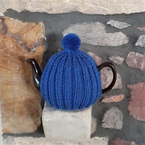 May include: A blue knitted teapot cosy with a pom-pom on top. The cosy is on a brown teapot.