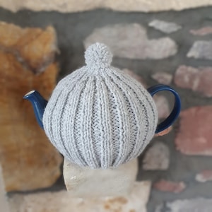 May include: A grey knitted teapot cosy with a pom-pom on top. The cosy is on a blue teapot.