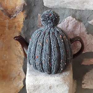 May include: A grey knitted teapot cosy with a pom-pom on top. The cosy has a speckled pattern with orange and white flecks.