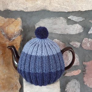 May include: A blue and grey knitted teapot cosy with a blue pom pom on top. The cosy is made of a soft, textured yarn and is designed to fit a standard-sized teapot.