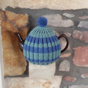 May include: A blue and green striped knitted teapot cosy with a pom pom on top. The cosy is on a brown teapot.