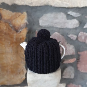 May include: A black knitted tea cosy with a pom-pom on top, covering a white teapot. The cosy has a ribbed texture and is set against a stone wall background.