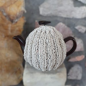 May include: A dark brown ceramic teapot encased in a light beige, vertically ribbed knitted cosy. The cosy covers the main body of the teapot, leaving the dark brown handle and spout exposed, and features a small dark brown knob on the lid.