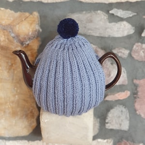 May include: A grey knitted teapot cosy with a blue pom pom on top. The cosy is on a brown teapot.