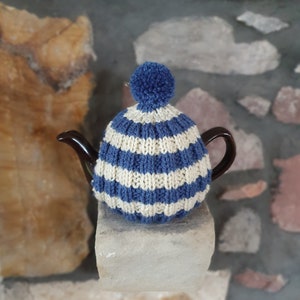 May include: A blue and white striped knitted teapot cosy with a pom pom on top. The cosy is on a stone surface.