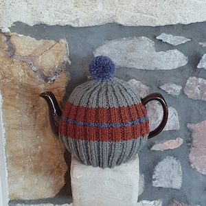 May include: A grey knitted teapot cosy with a brown and blue striped pattern and a blue pom pom on top.