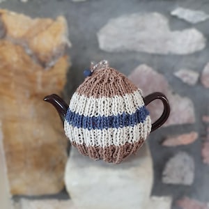 May include: A dark brown ceramic teapot covered by a hand-knitted tea cosy. The cosy features horizontal stripes in shades of brown, cream, and blue, with a ribbed texture and a small blue bead accent at the top centre of the rounded cover.