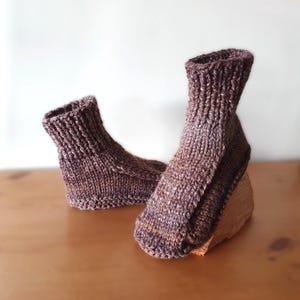 Könnte beinhalten: Ein Paar handgestrickte Socken in Braun- und Lilatönen. Die Socken haben einen gerippten Bund und ein strukturiertes Strickmuster. Sie liegen auf einer Holzoberfläche, was ihre warme, handgefertigte Qualität unterstreicht.