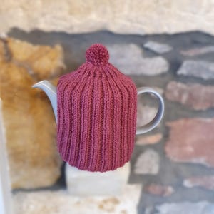 Könnte beinhalten: Ein gestrickter Teewärmer in tiefem Rosé, der eine weiße Teekanne bedeckt. Der Teewärmer hat eine gerippte Textur und einen kleinen Bommel oben. Die Teekanne steht vor einem verschwommenen Hintergrund aus Stein und Ziegeln.