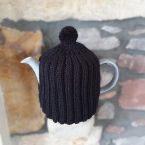 May include: Black knitted teapot cosy with a pom pom on top. The cosy is on a grey teapot.