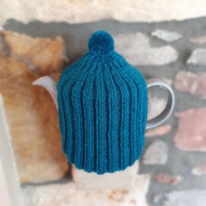 Peut inclure: Une housse de théière en tricot bleu sarcelle avec un pompon sur le dessus. La housse est sur une théière grise.
