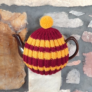 May include: A burgundy and yellow striped knitted tea cosy with a yellow pom-pom. The cosy is covering a brown teapot. The tea cosy is made of a ribbed knit fabric. The teapot is on a stone surface.