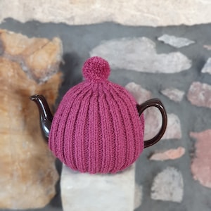 May include: A pink knitted teapot cosy with a pom pom on top. The cosy is on a brown teapot.